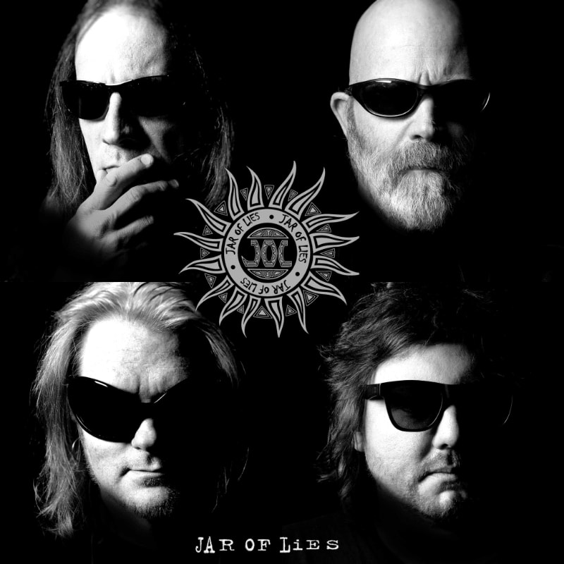 Four men wearing sunglasses are arranged in a square with a circular logo in the center against a black background.