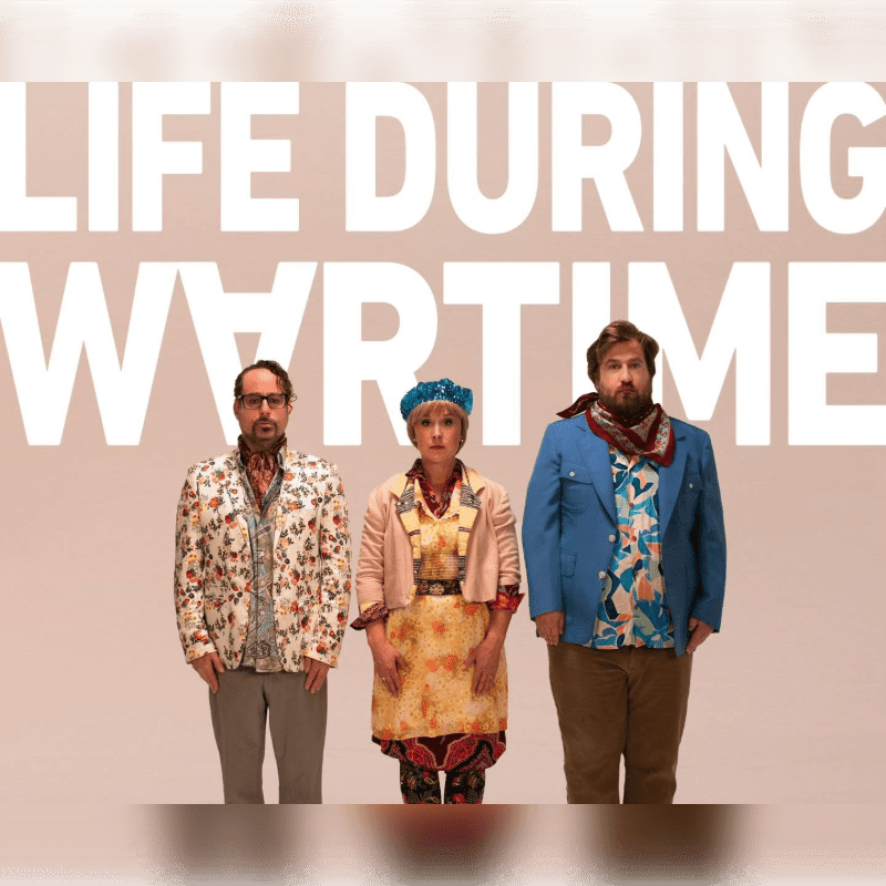 Three people in colorful outfits stand in front of a pale pink background with large white text that reads "LIFE DURING WARTIME."