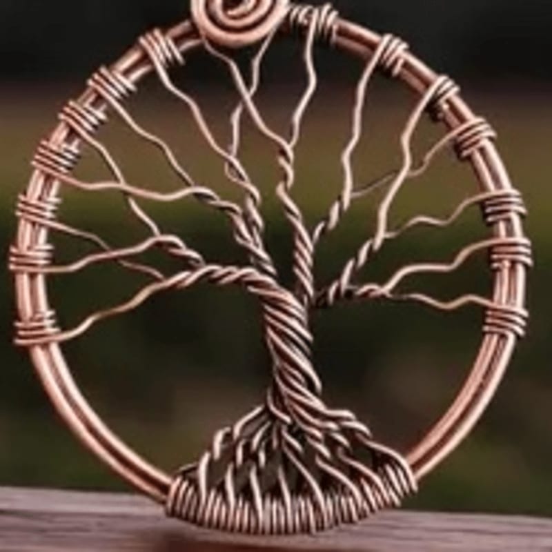 A copper wire tree of life pendant with intricate branches and roots is set against a blurred green and brown background.