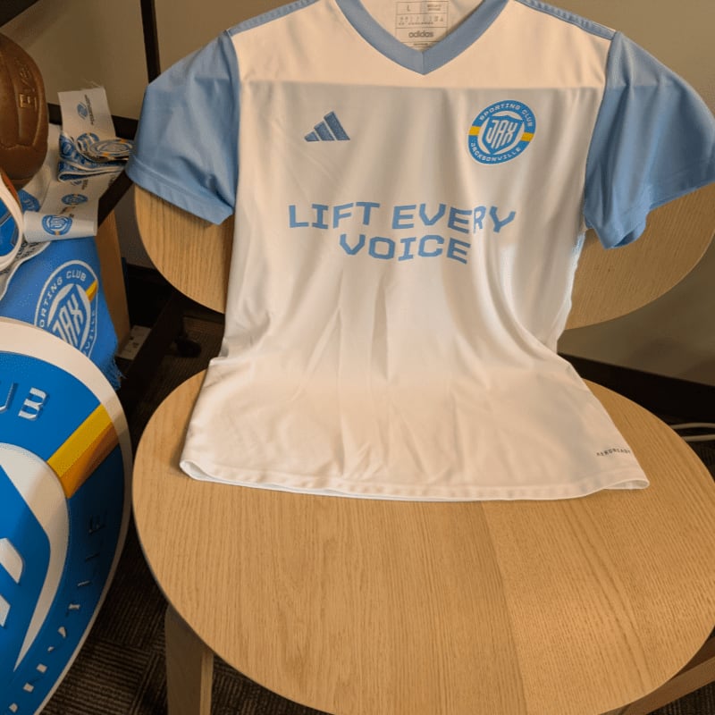 A white and light blue soccer jersey with the slogan "LIFT EVERY VOICE" is displayed on a wooden chair, with other team merchandise visible in the foreground and a blurred background.