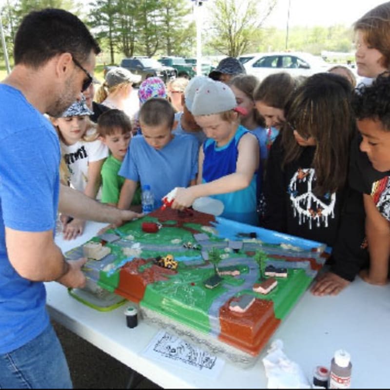 A man in a blue shirt demonstrates a miniature town model to a group of children in the foreground, with cars and trees on the model, while a blurred background shows more people and parked vehicles.