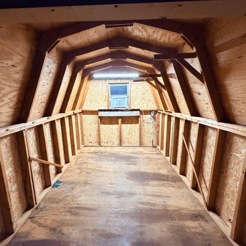 The wooden floor of an empty shed leads to a small window with a light above it, framed by the shed's arched roof and walls.