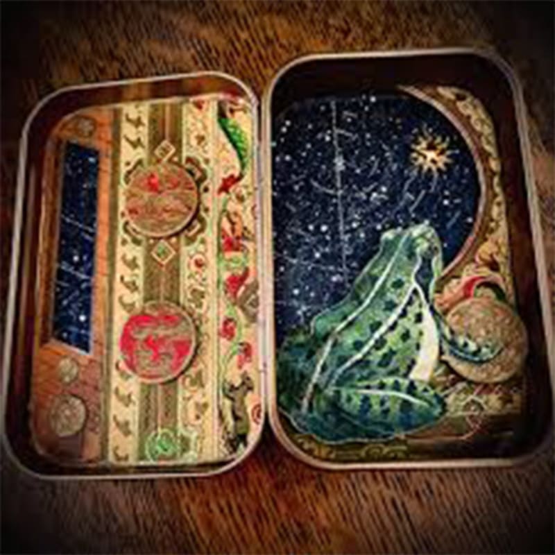 A green frog sits on a starry night sky within an open tin, with the other half of the tin decorated with ornate patterns and celestial motifs.