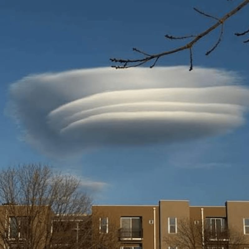A large, layered lenticular cloud hovers over a modern apartment building with bare trees in the foreground.