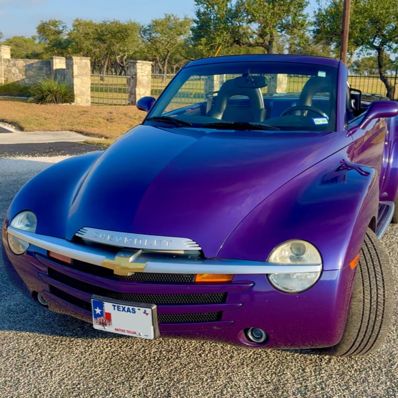 A vibrant purple Chevrolet SSR convertible is parked on a gravel driveway with a stone wall and trees in the background.