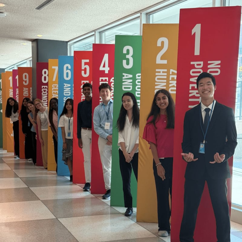 A group of young people stand in front of colorful banners representing the UN Sustainable Development Goals, with a man in a suit gesturing towards the camera in the foreground.