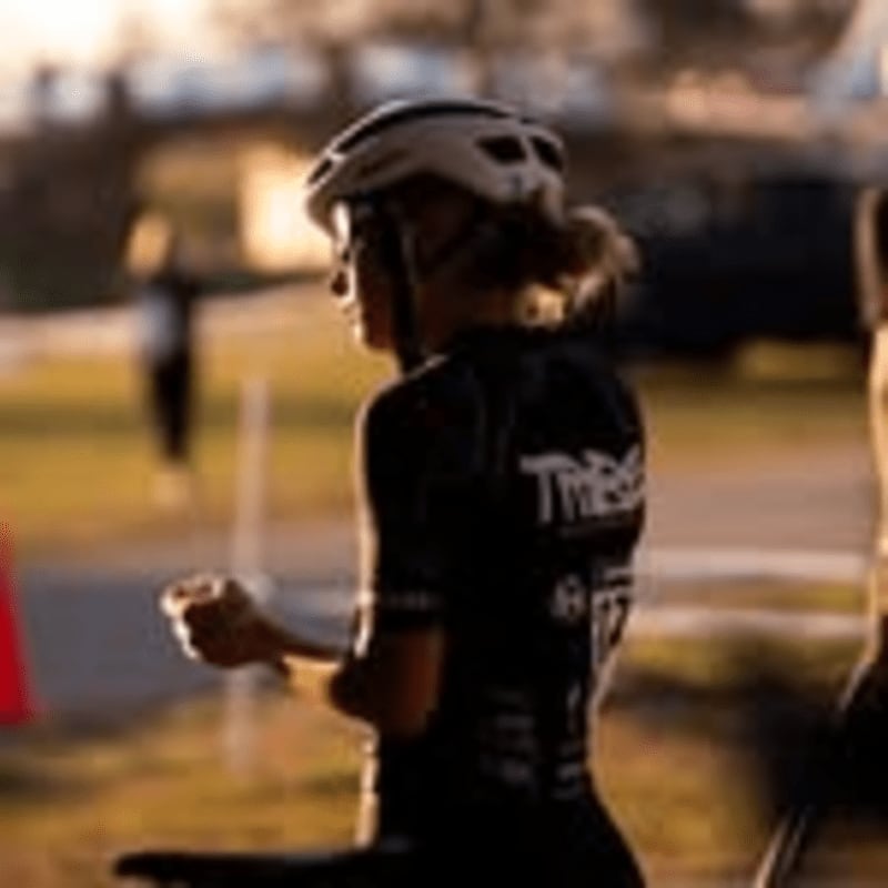 A cyclist in a helmet and racing attire stands in the foreground with a blurred outdoor background.