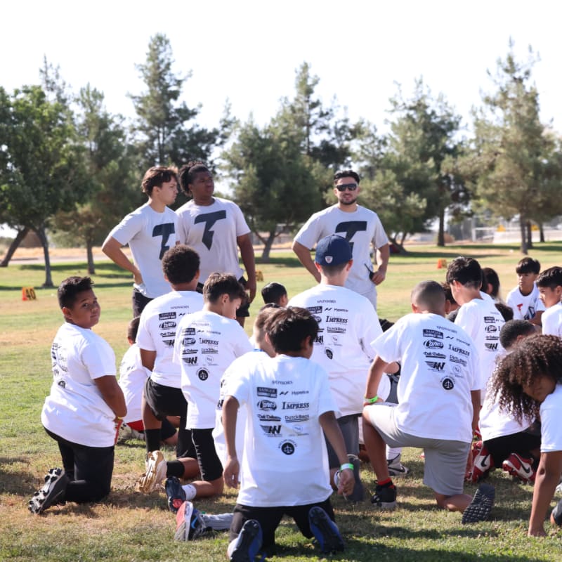 A group of young boys in white t-shirts are gathered on a grassy field, with older teens and adults in the background under a clear sky with trees.