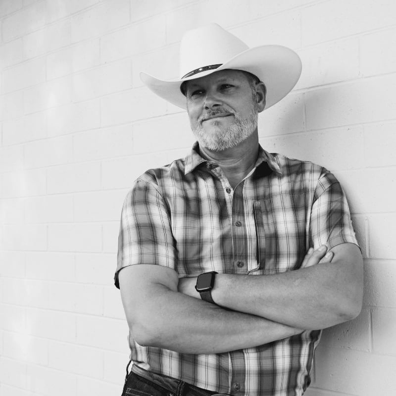 A man in a cowboy hat and plaid shirt leans against a white brick wall with his arms crossed.