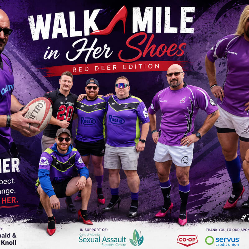 A group of men in purple rugby shirts and shorts, some wearing high heels, pose for a photo in front of a "Walk Mile in Her Shoes" banner.