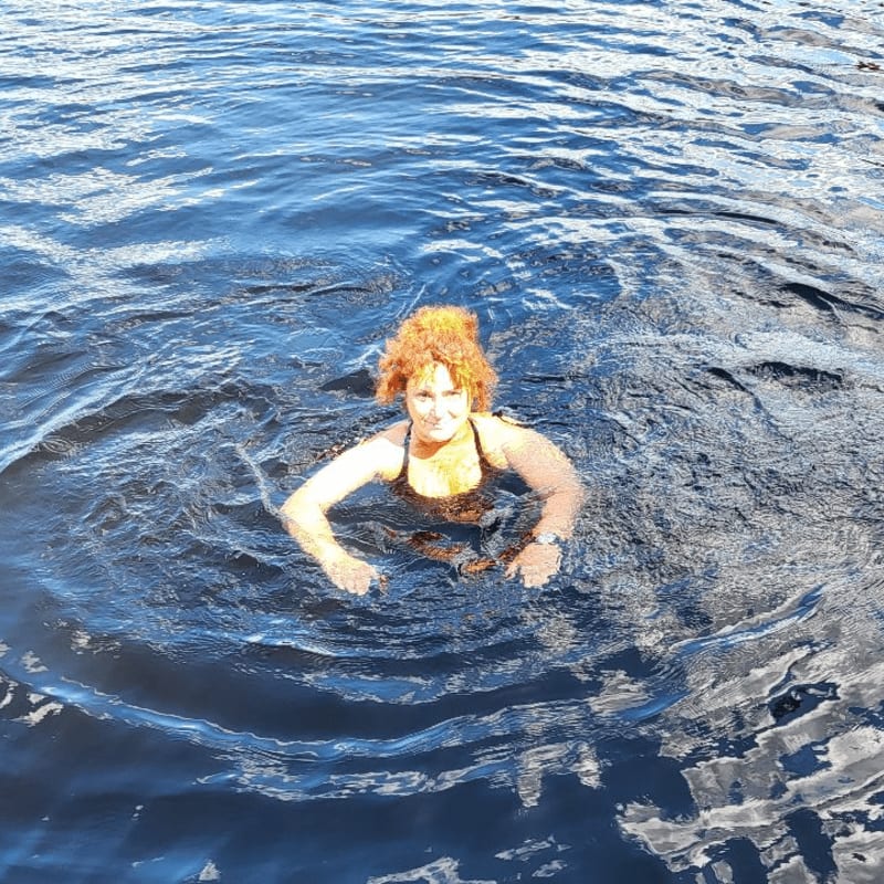 A woman with curly red hair swims in dark blue water, with ripples extending to the edges of the frame.