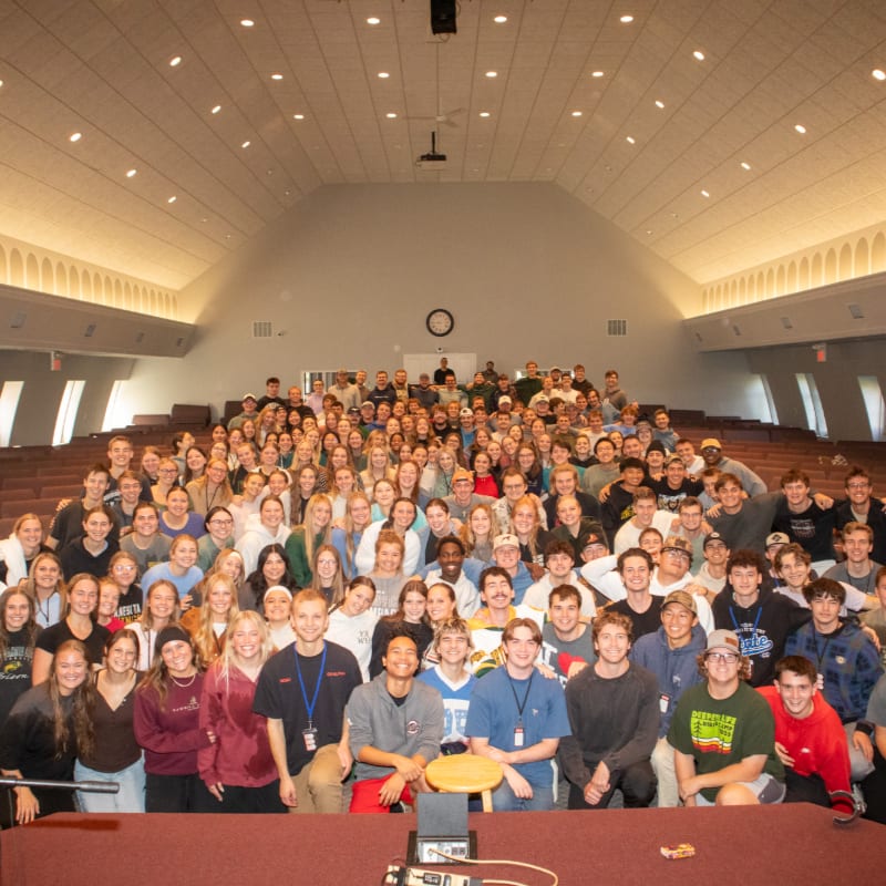 A large group of young people are gathered in a lecture hall with tiered seating.