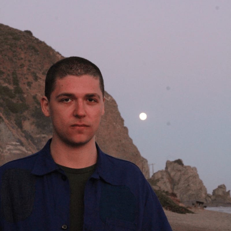 A young man stands on a beach with a rocky, moonlit coastline behind him.