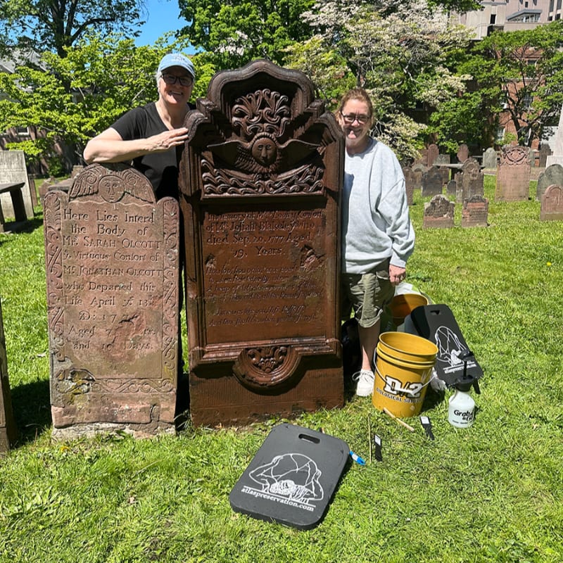 Two women stand in a grassy cemetery next to a large, ornate headstone and a smaller, weathered one, with cleaning supplies scattered on the ground.