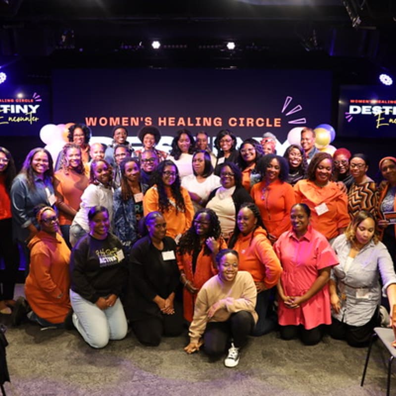 A diverse group of women in casual attire pose for a photo in front of a stage with "WOMEN'S HEALING CIRCLE" displayed on a screen.