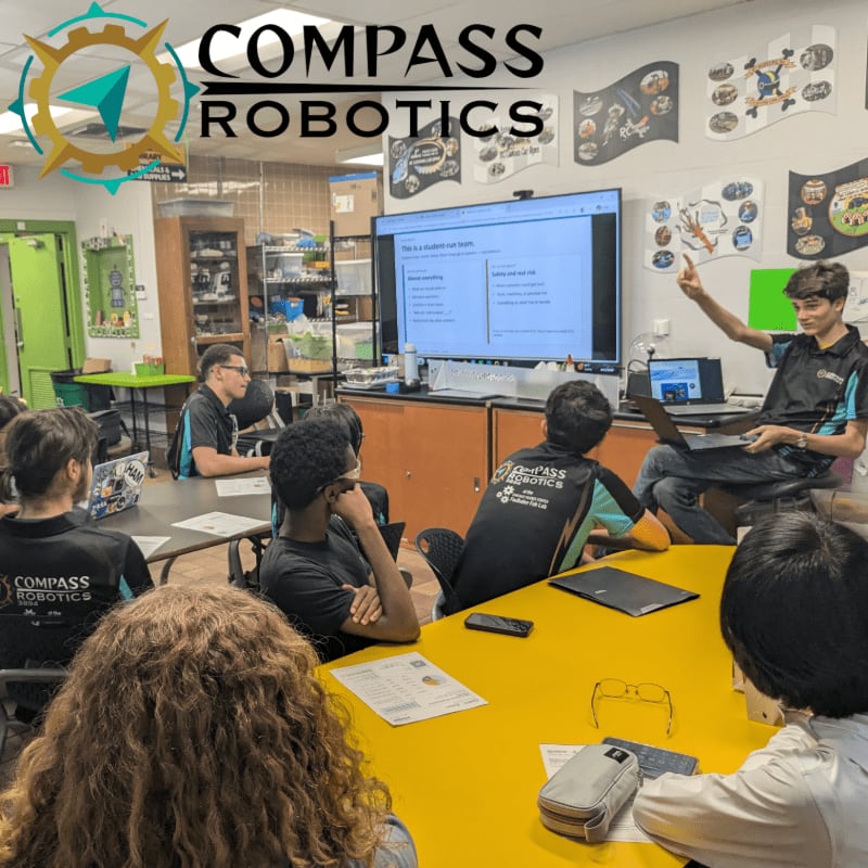 Students in the foreground are seated around a yellow table, facing a large screen in the background where a presenter is pointing, with the "COMPASS ROBOTICS" logo visible at the top.
