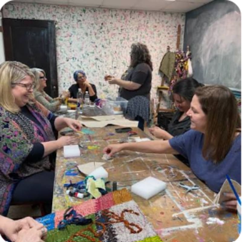 A group of women are gathered around a craft table in a room with a colorful, textured wall.