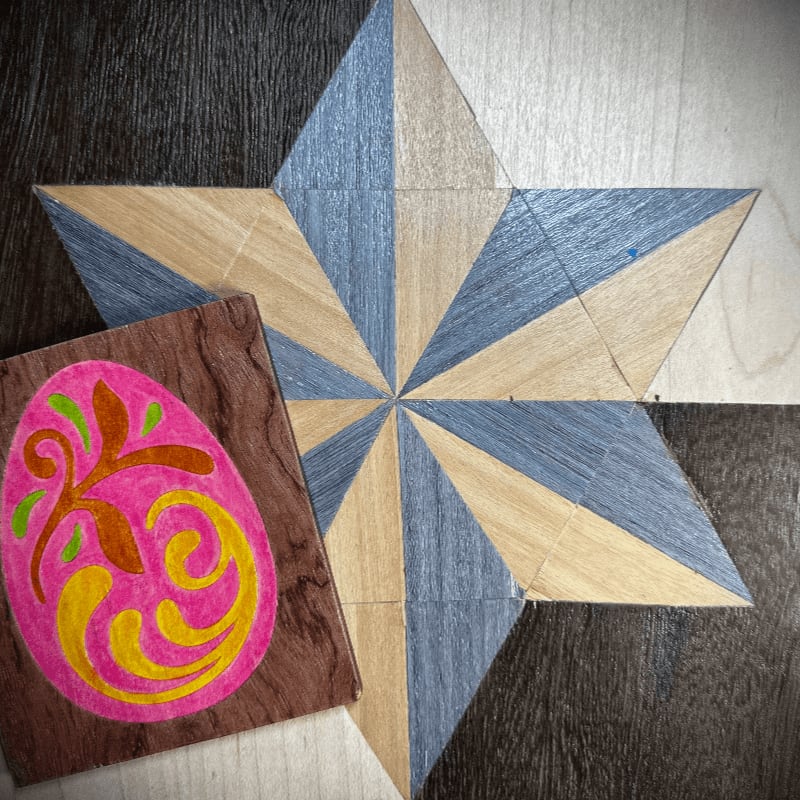 A pink, yellow, and green decorated egg sits on a wooden block in front of a large, multi-colored starburst pattern on a floor.