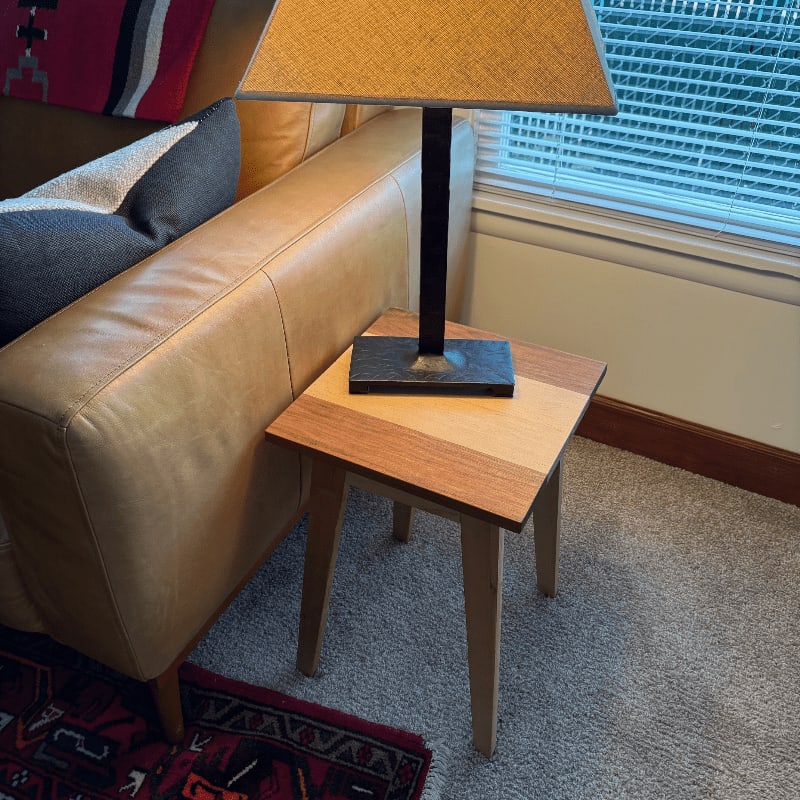 A small wooden side table with a lamp on it sits next to a tan leather couch with patterned pillows, in front of a window with blinds.