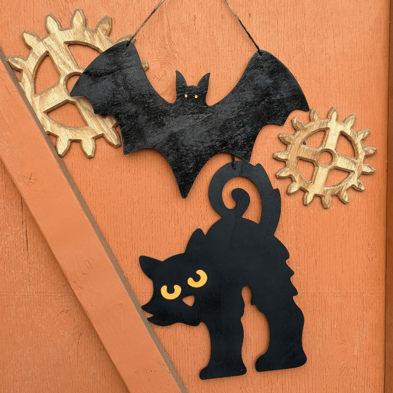 A black bat and a black cat with yellow eyes hang in front of two golden gears against an orange wooden background.