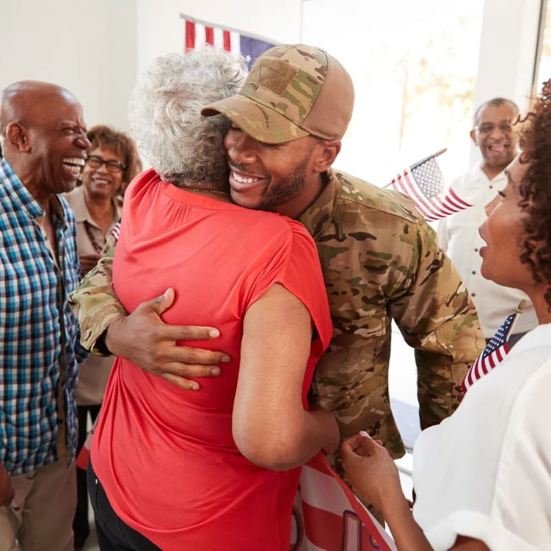 A soldier in uniform embraces an older woman in the foreground, while a group of smiling family members and American flags are visible in the background.