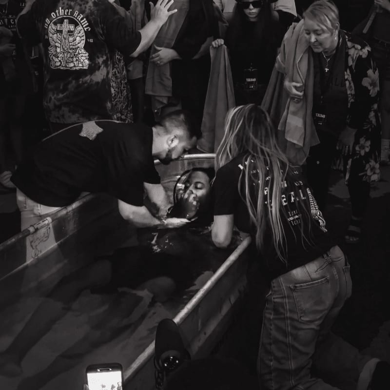 In the foreground, a person is being baptized in a shallow pool by two individuals, while a crowd of onlookers stands in the background.