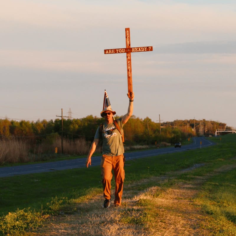 A man in a conical hat walks along a grassy path carrying a cross that reads "ARE YOU READY?", with a road and trees in the background under a pale sky.