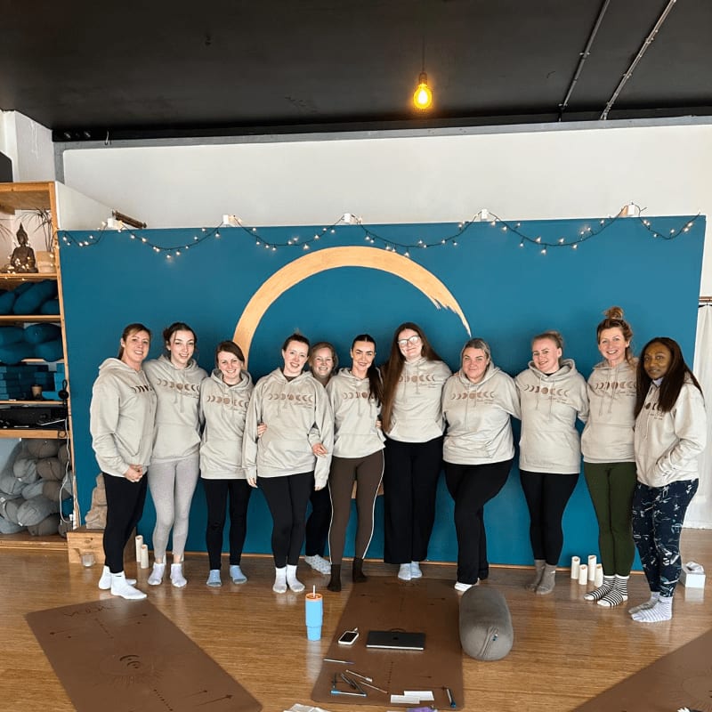 A group of women in matching hoodies stand in front of a teal backdrop with a crescent moon design, while yoga mats and props are visible in the foreground.
