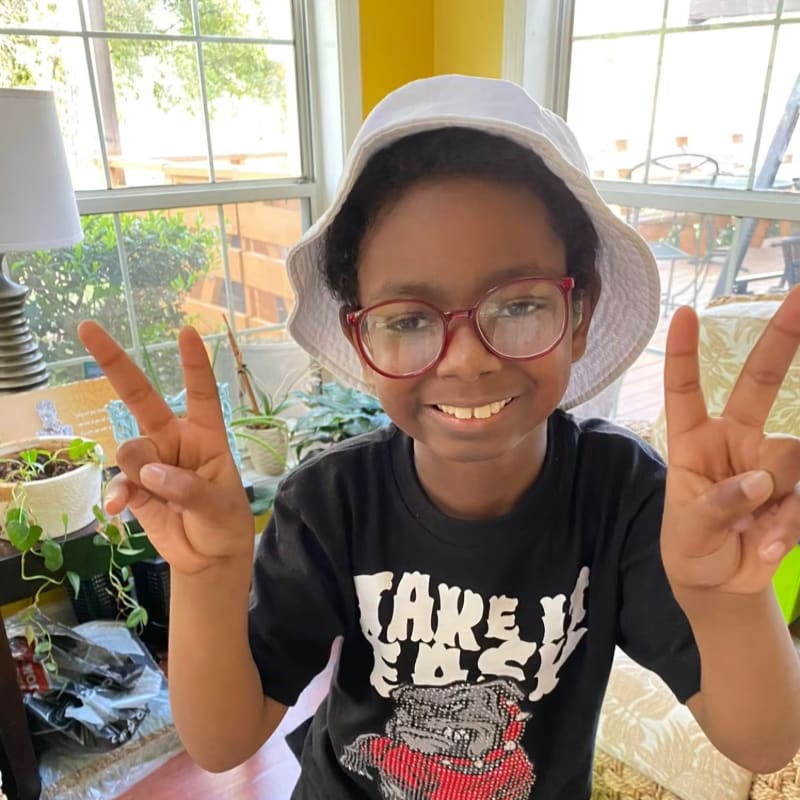 A young boy in a black t-shirt and white bucket hat smiles and holds up peace signs in the foreground, with a sunlit room and plants visible in the background.