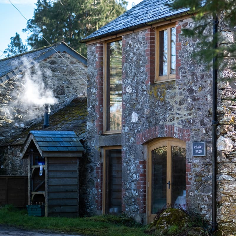 A rustic stone barn with a smoking chimney stands in the foreground, while a wooded area is visible in the background.