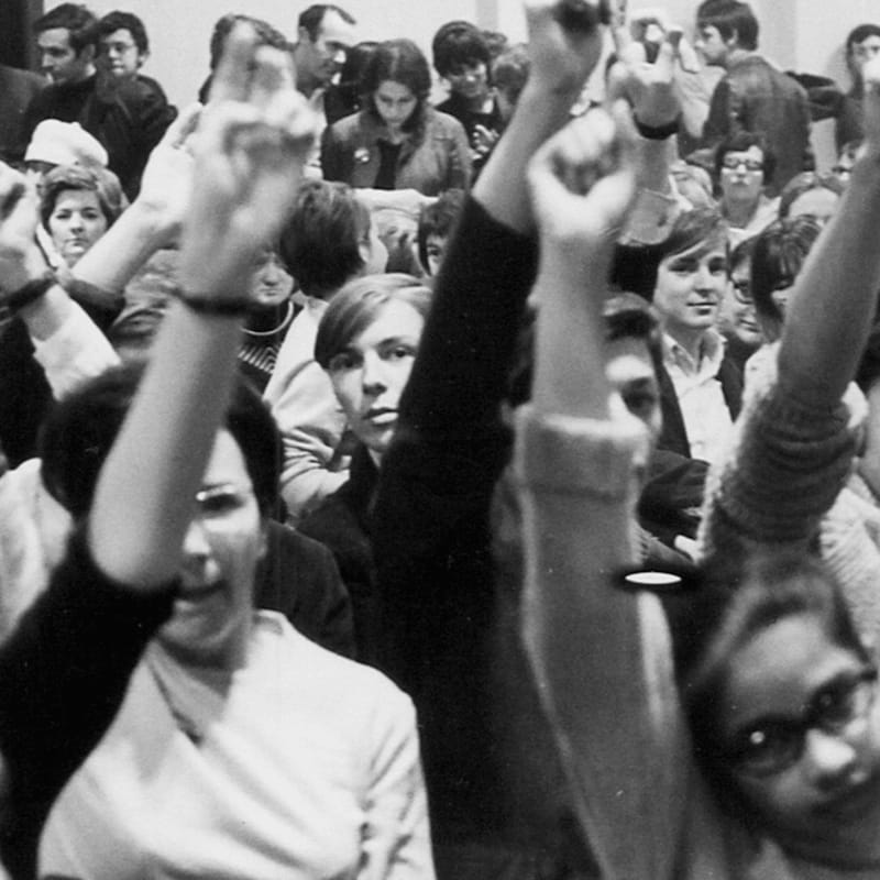 In the foreground, several people raise their hands in a crowd, while the background shows more faces looking forward.