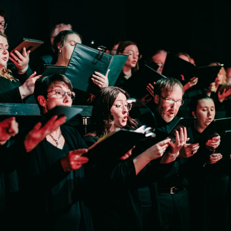 A choir of singers in black attire, holding sheet music, performs on a dimly lit stage.