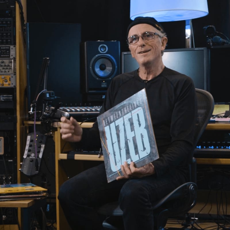 A smiling man in a black beanie holds a record album in a recording studio filled with audio equipment.