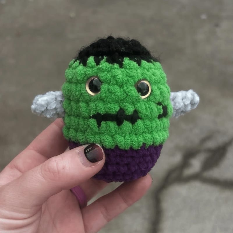 A crocheted Frankenstein's monster toy with black hair, green body, and purple pants is held in a hand with dark nail polish against a blurred concrete background.