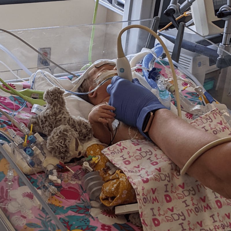 A gloved hand holds a medical device over a baby in an incubator, surrounded by medical equipment and a stuffed animal.