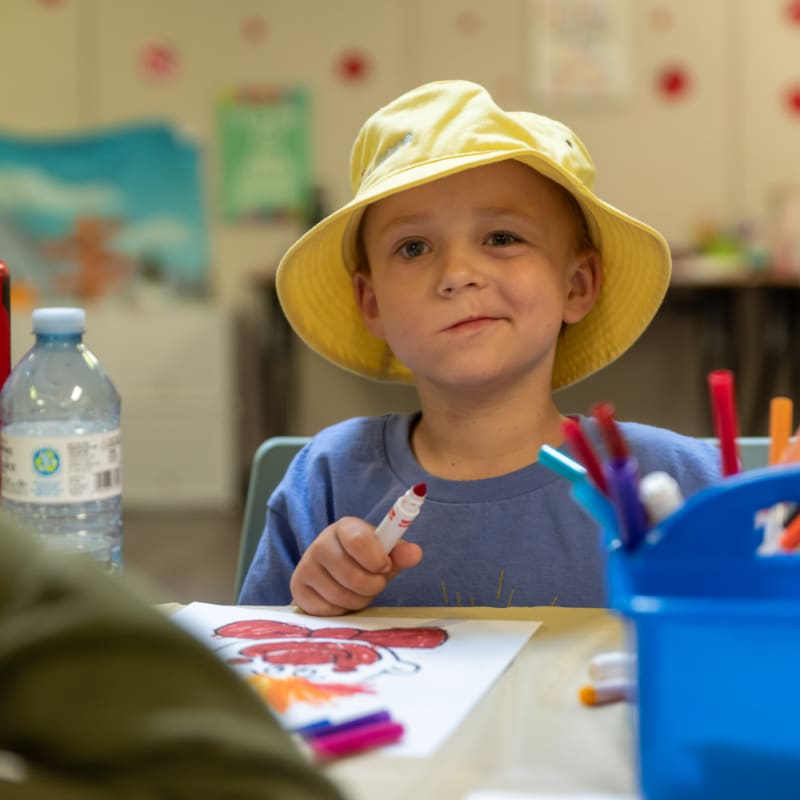 A young boy in a yellow hat smiles while coloring a picture, with a water bottle and art supplies on the table and a blurred classroom in the background.
