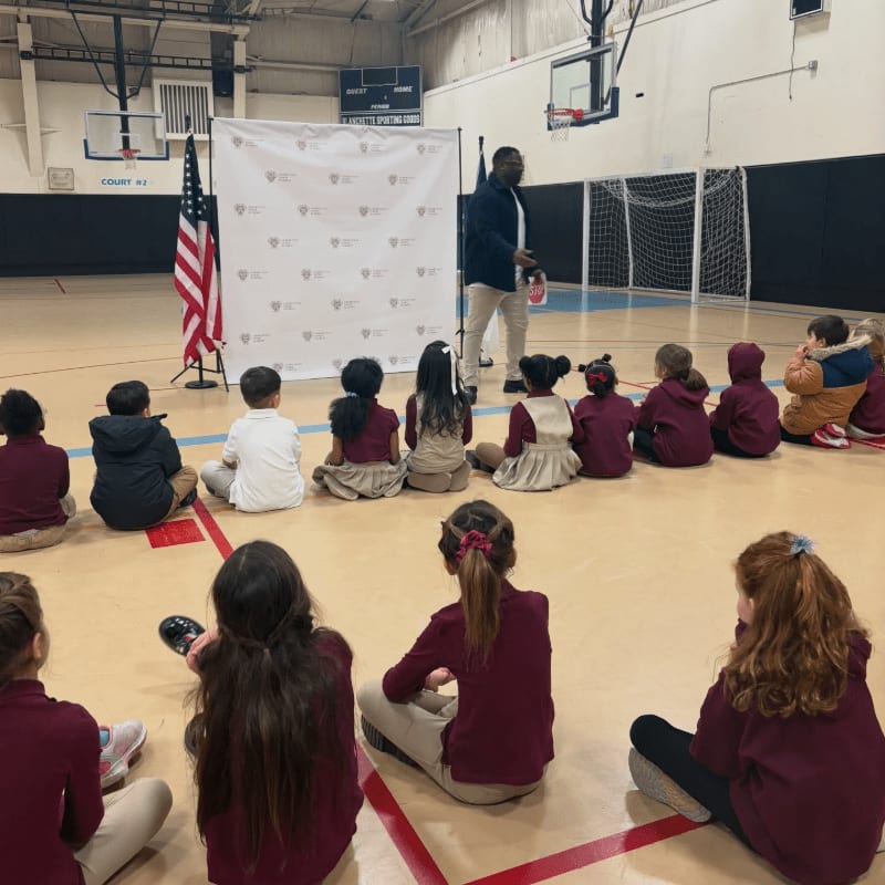 Children in maroon uniforms sit on a gymnasium floor in the foreground, facing a man standing before a backdrop with an American flag in the background.