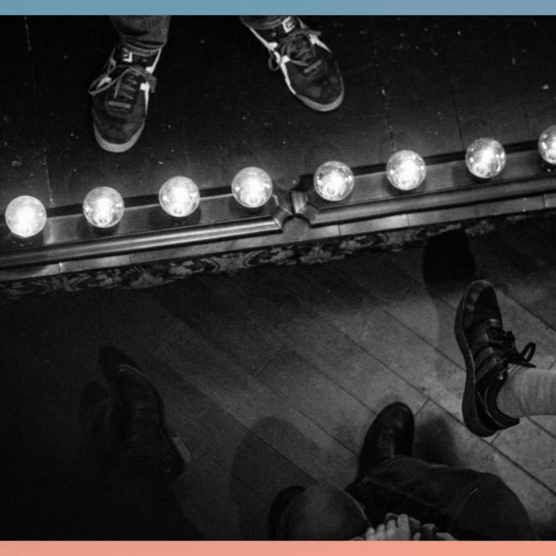Feet in sneakers stand on a wooden stage in front of a row of illuminated bulbs, with more feet visible in the foreground.