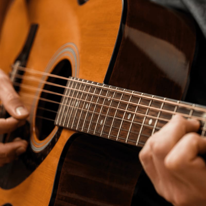 Hands strumming the strings of a warm-toned acoustic guitar against a dark, blurred background.