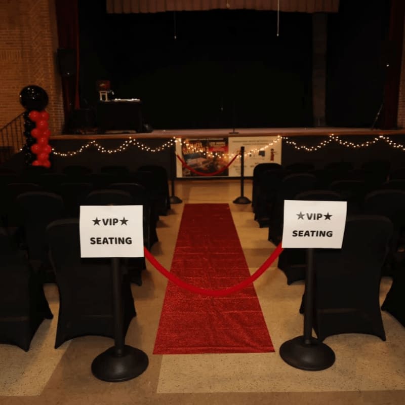 A red carpet leads to a stage with black chairs arranged in rows, framed by balloon decorations and a brick wall.
