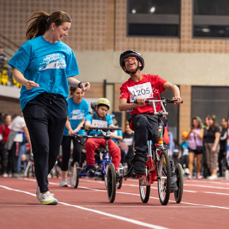 A woman in a blue shirt runs alongside a boy on a red tricycle in the foreground, while other participants and spectators fill the blurred background of an indoor track.