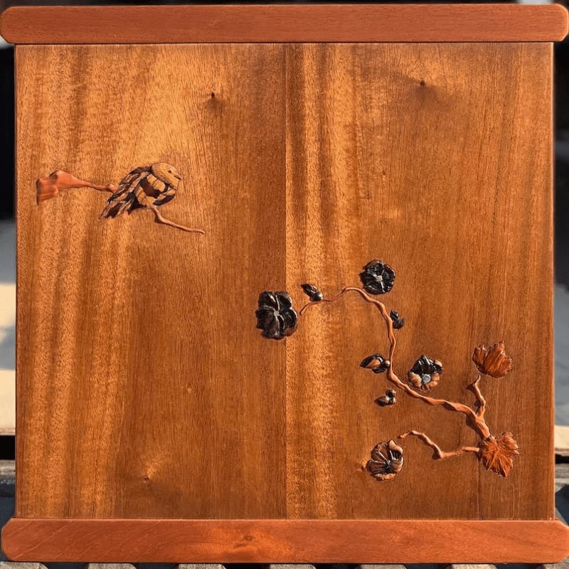 A carved wooden panel features a bird on a branch in the foreground against a warm, grained wood background.