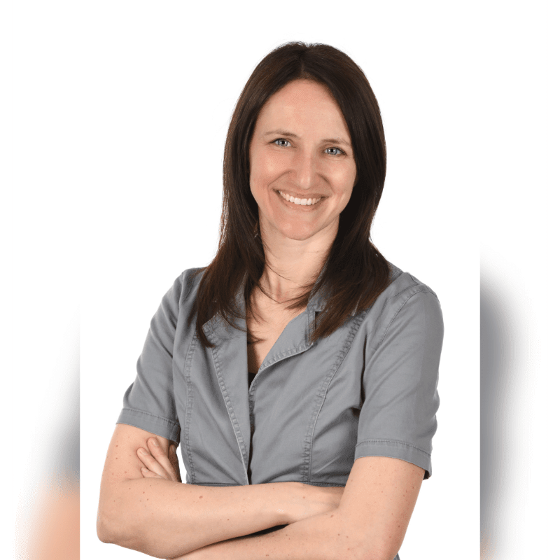 A smiling woman with dark hair and a gray shirt stands with her arms crossed against a plain white background.