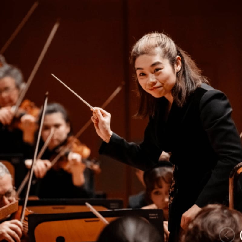 A female conductor leads an orchestra, with musicians playing their instruments in the blurred background.