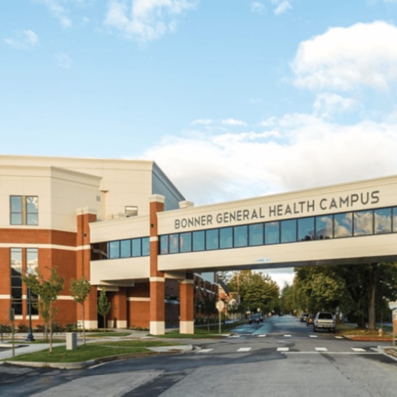 A modern brick building with a skybridge labeled "BONNER GENERAL HEALTH CAMPUS" spans over a street lined with trees under a partly cloudy sky.