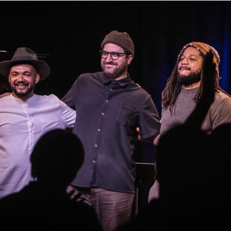 Three men smile at the camera in the foreground, with a dark, blurred background suggesting a performance space.