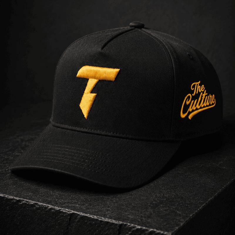 A black baseball cap with a yellow "T" logo and "The Culture" embroidered on the side rests on a dark, textured surface against a black background.