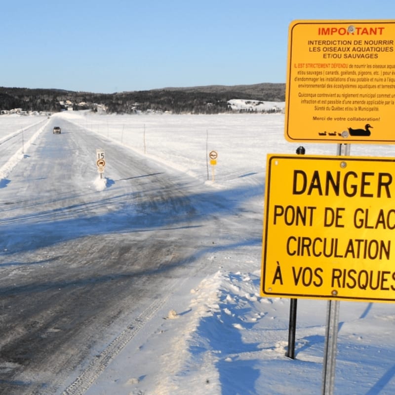 Yellow danger signs stand in the foreground next to a snow-covered road leading to a distant, treelined horizon under a clear blue sky.