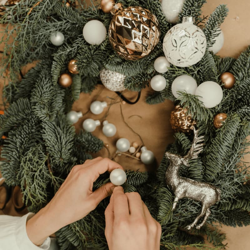 Hands arrange white ornaments on a lush green Christmas wreath adorned with gold and silver baubles and a reindeer figurine, set against a plain brown background.