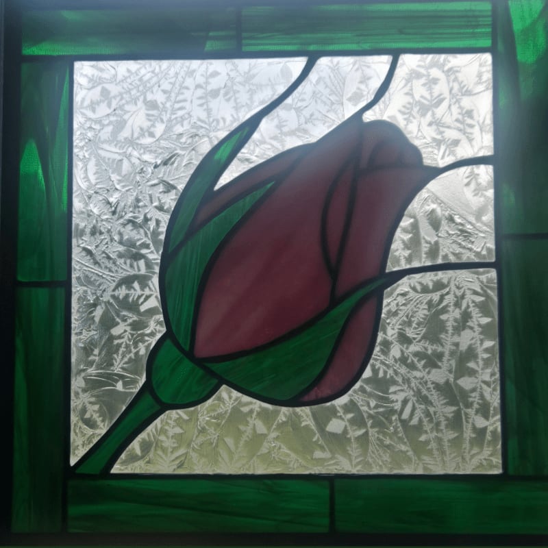 A stained glass rosebud with green leaves is set against a frosted glass background with a leafy pattern, all framed by green stained glass.